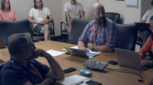 Three people sit around a conference table, one facing away from the camera and two on the other side of the table facing toward the camera. There are several people in the background as well. There are computers and a phone on the conference table.